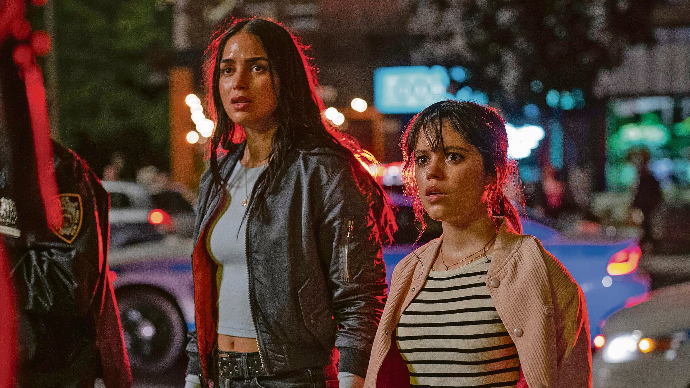 Melissa Barrera y Jenna Ortega de pie en una carretera. Miran con cara medio de sopresa o terror.